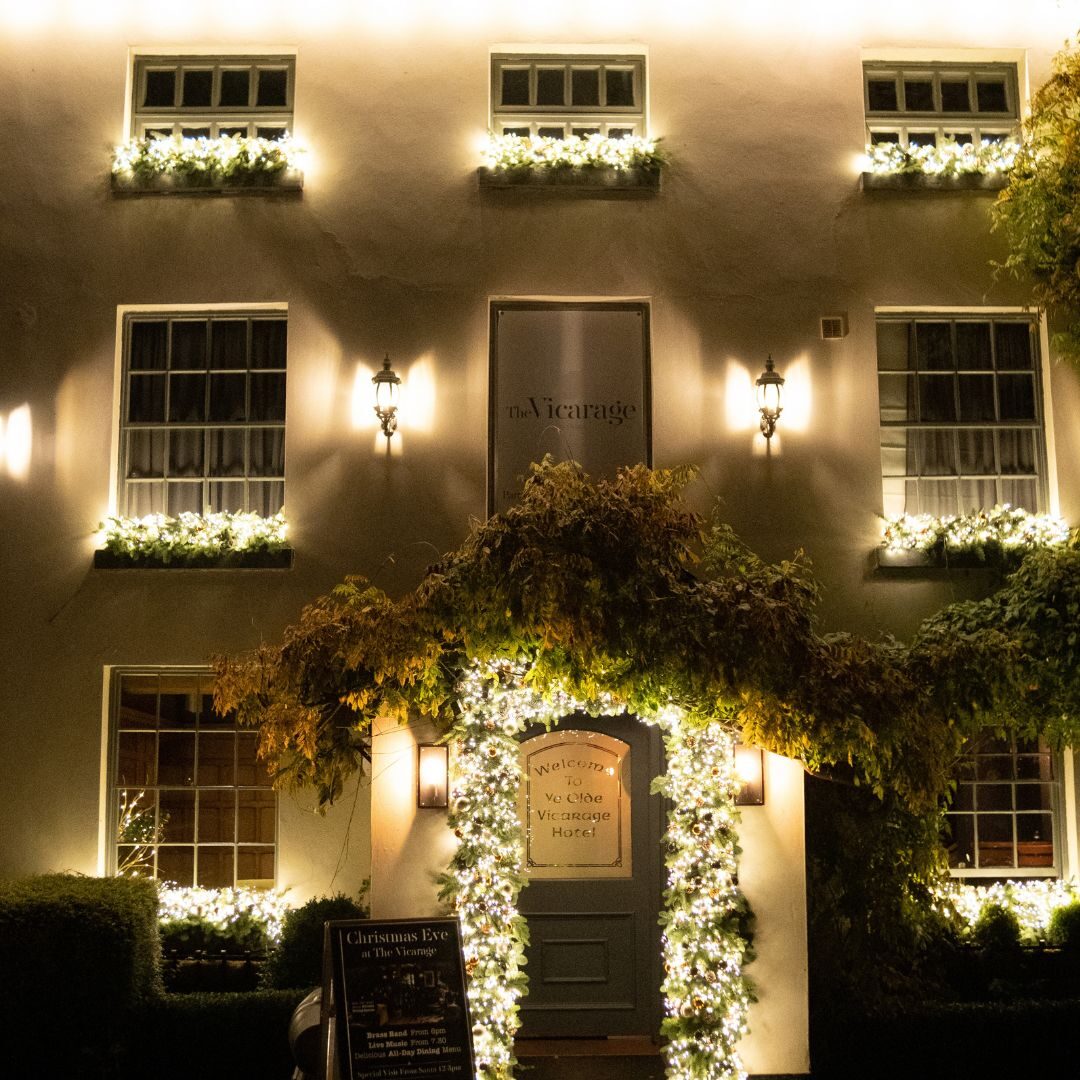 The Old Vicarage at night, its white front lit by wall lamps and an ivy-framed doorway strung with lights